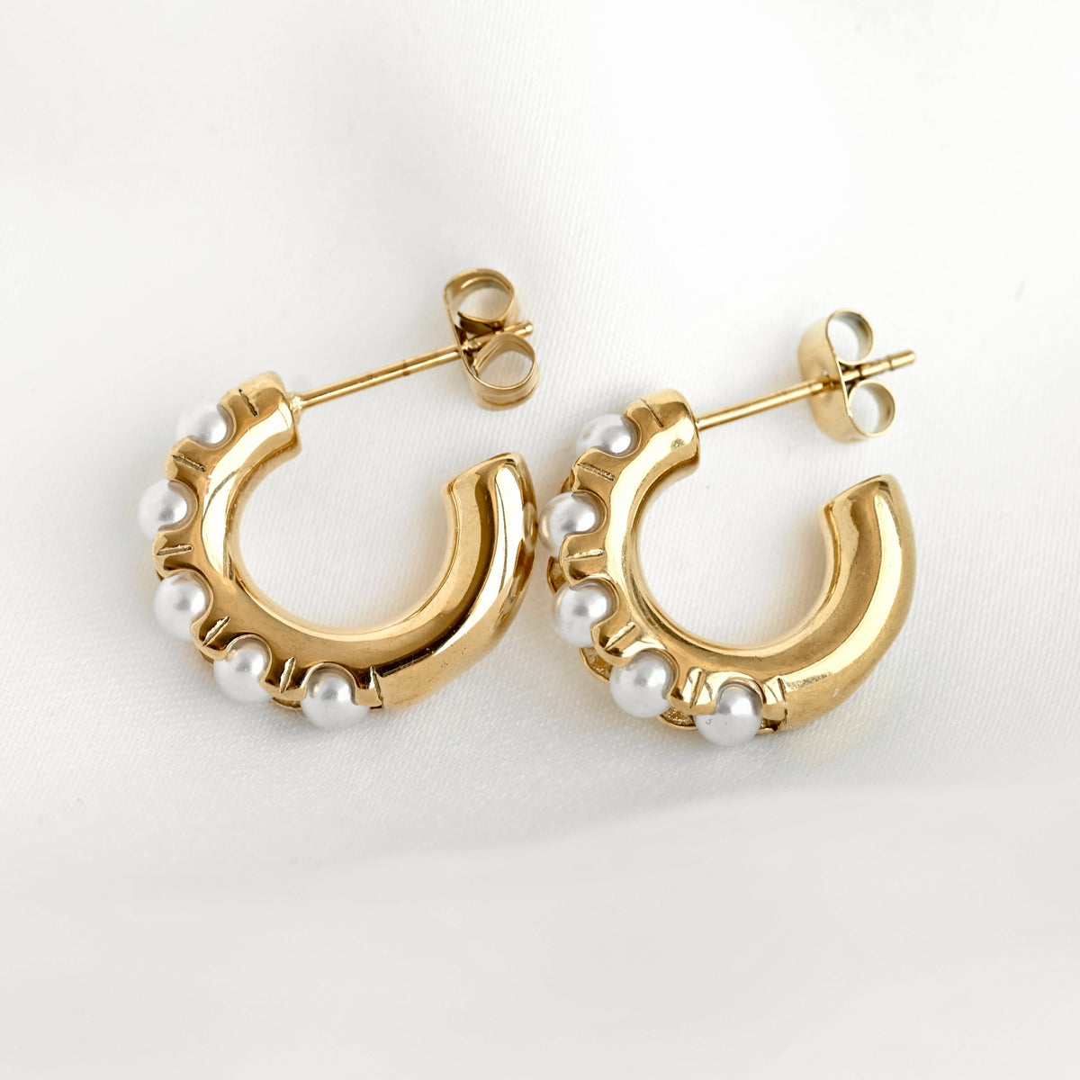 A pair of gold C-shaped hoop earrings decorated with small white pearls along the front edge, displayed on a clean white fabric background.
