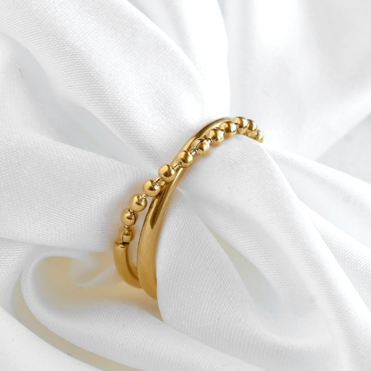 A gold beaded chain criss cross ring with a ball chain crossing oer a smooth gold band on a white draped fabric background.