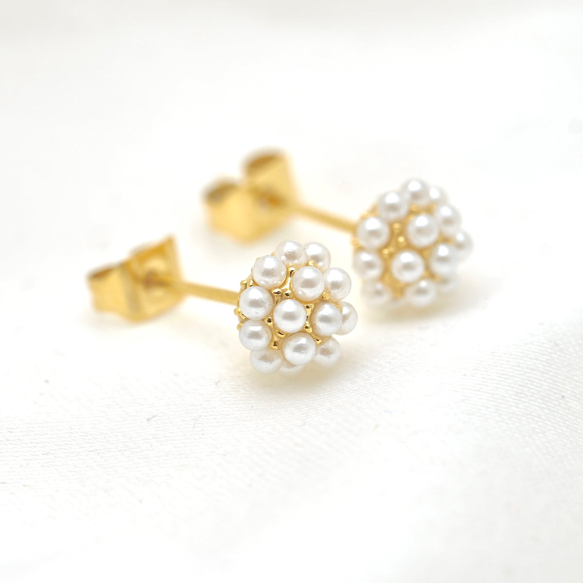 A pair of gold stud earrings with white pearl flower clusters, displayed on a clean white textured fabric background.