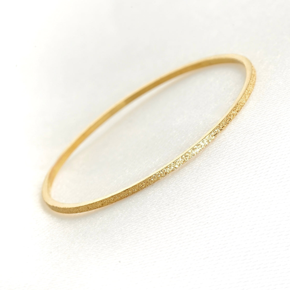 A delicate, thin gold bandwith a textured, glittery finish, displayed on a clean white textured background.