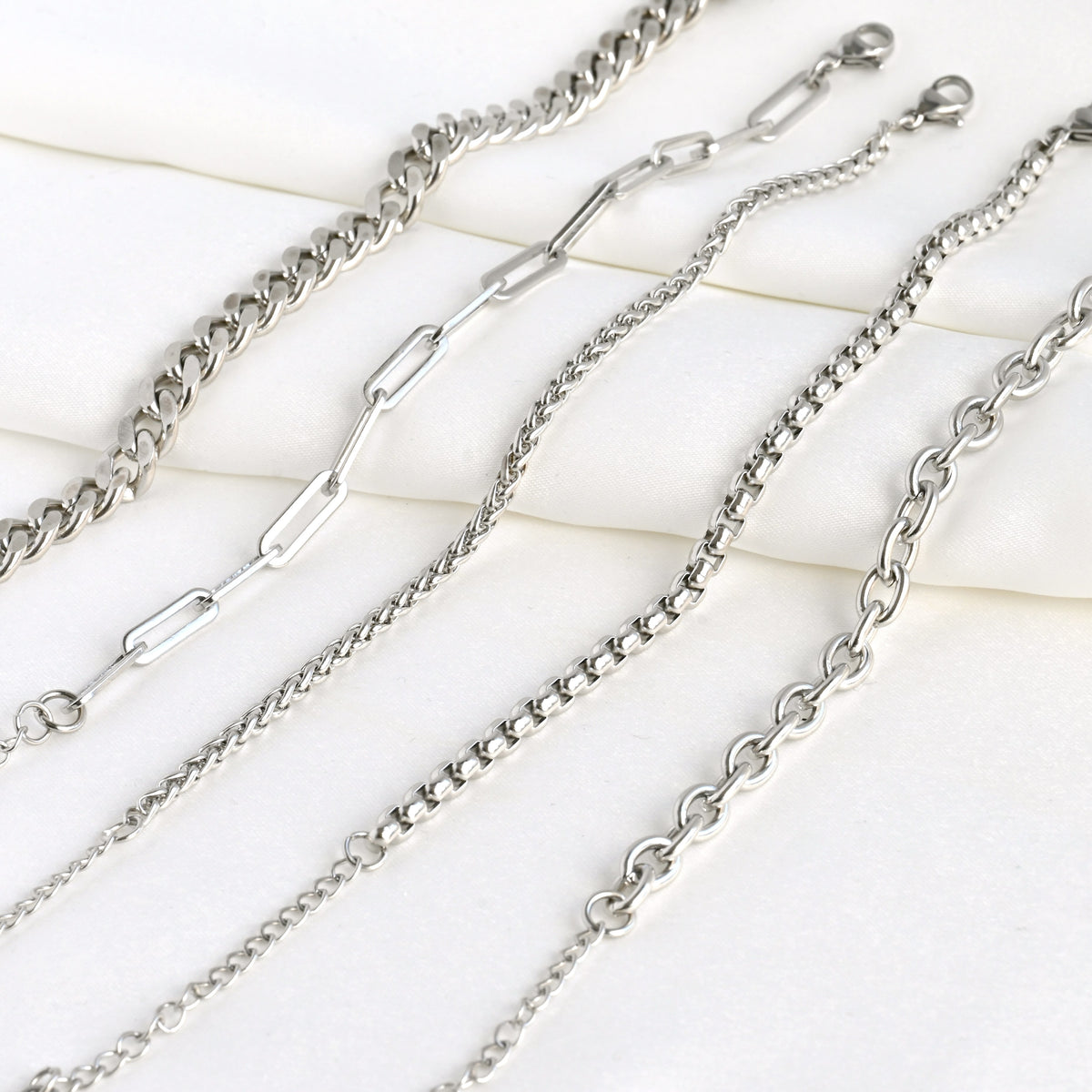 Silver chain bracelets on a light satin fabric. From left to right: Curb, paperclip, wheat, rounded box and rolo