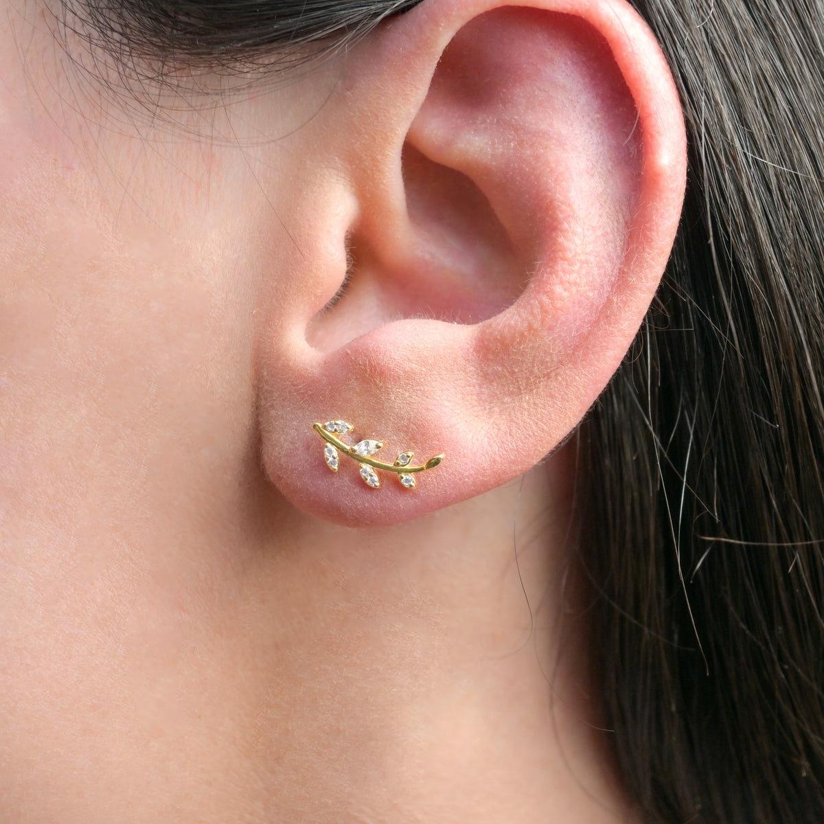 A close-up image of a woman's ear wearing a gold olive branch stud earring with cubic zirconia stones.