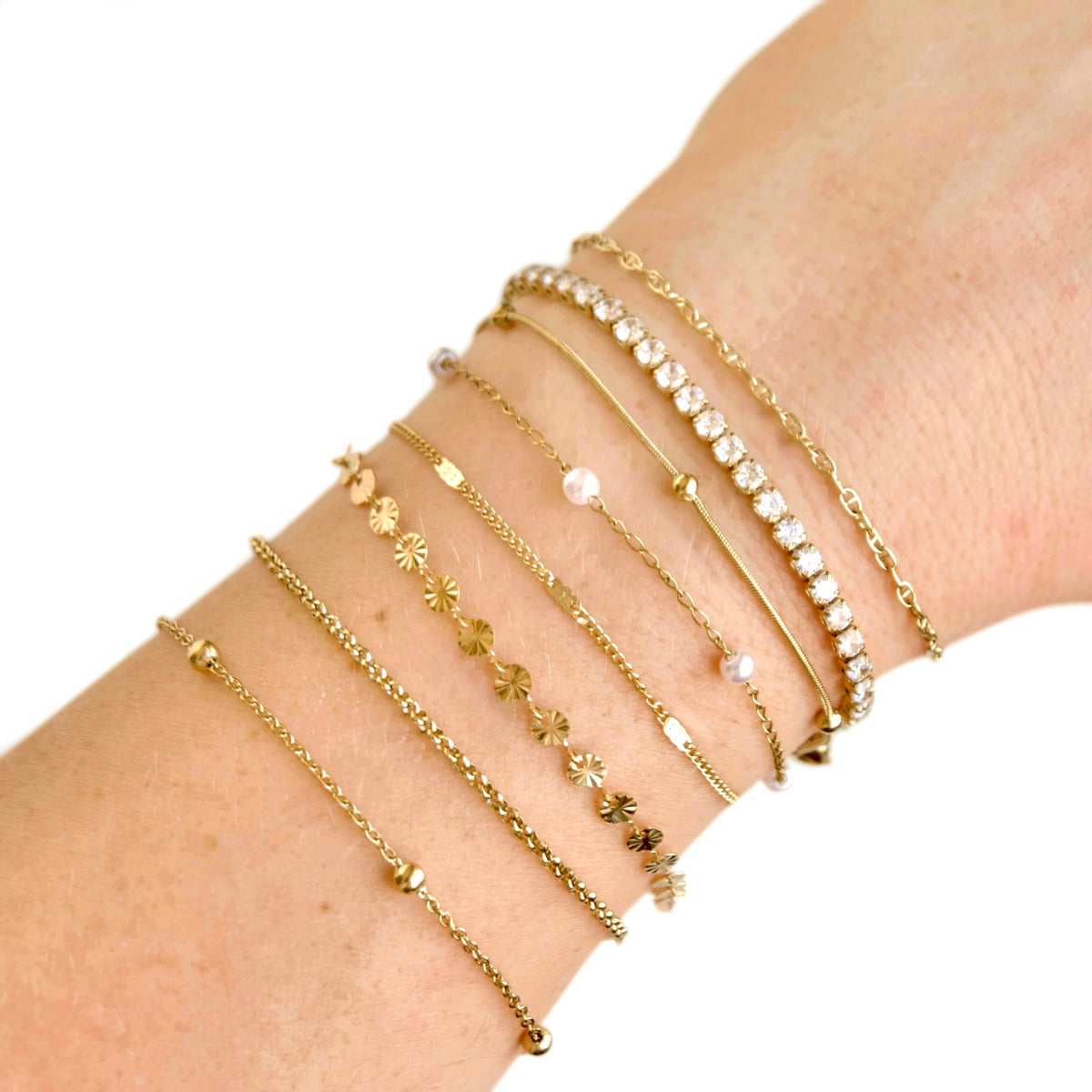 A close-up of a person's wrist wearing a stack of delicate gold bracelets, including floral, pearl, and engraved names like 'SUNRISE' and 'PEARL'.