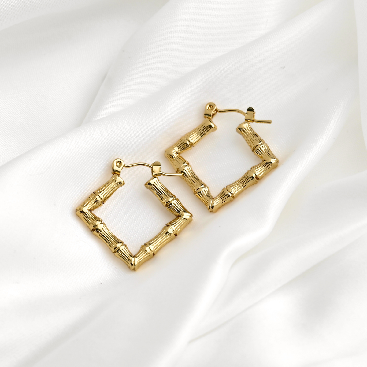 A pair of gold square hoop earrings designed to look like bamboo stalks, resting on a draped white satin fabric background.