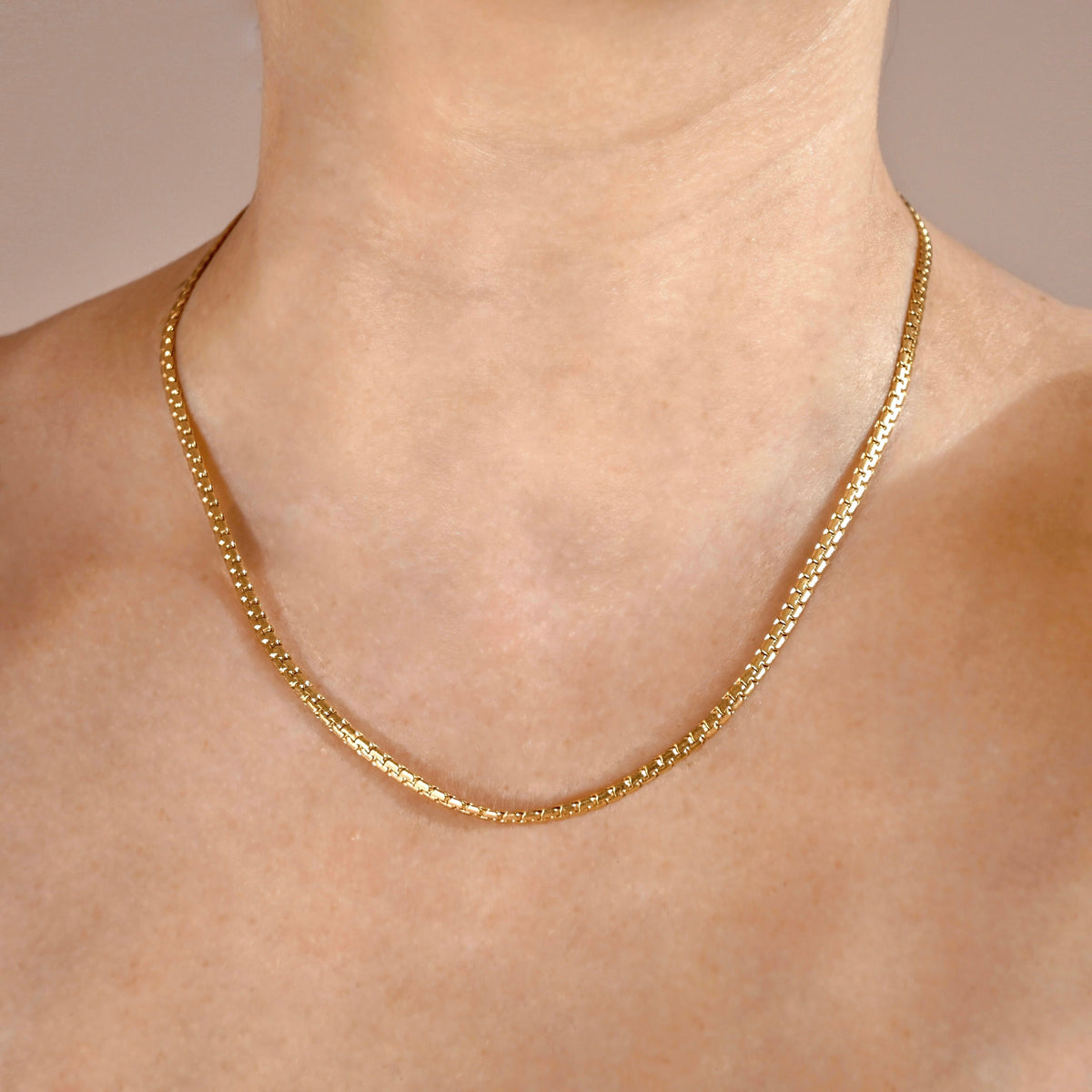 A close-up of a person's neck and upper chest wearing a thick gold snake chain necklace against a soft beige background.