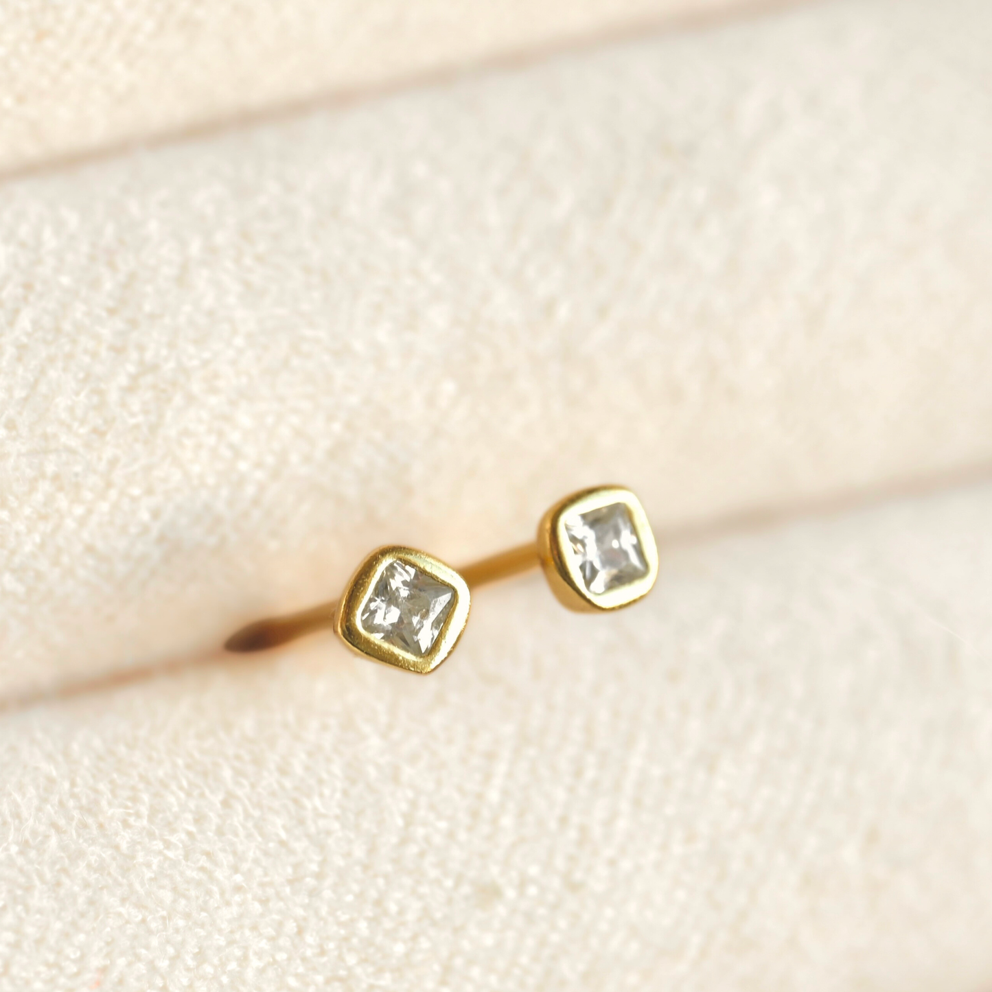 A pair of gold stud earrings with square-cut clear stones, displayed on a textured cream-colored jewelry box.