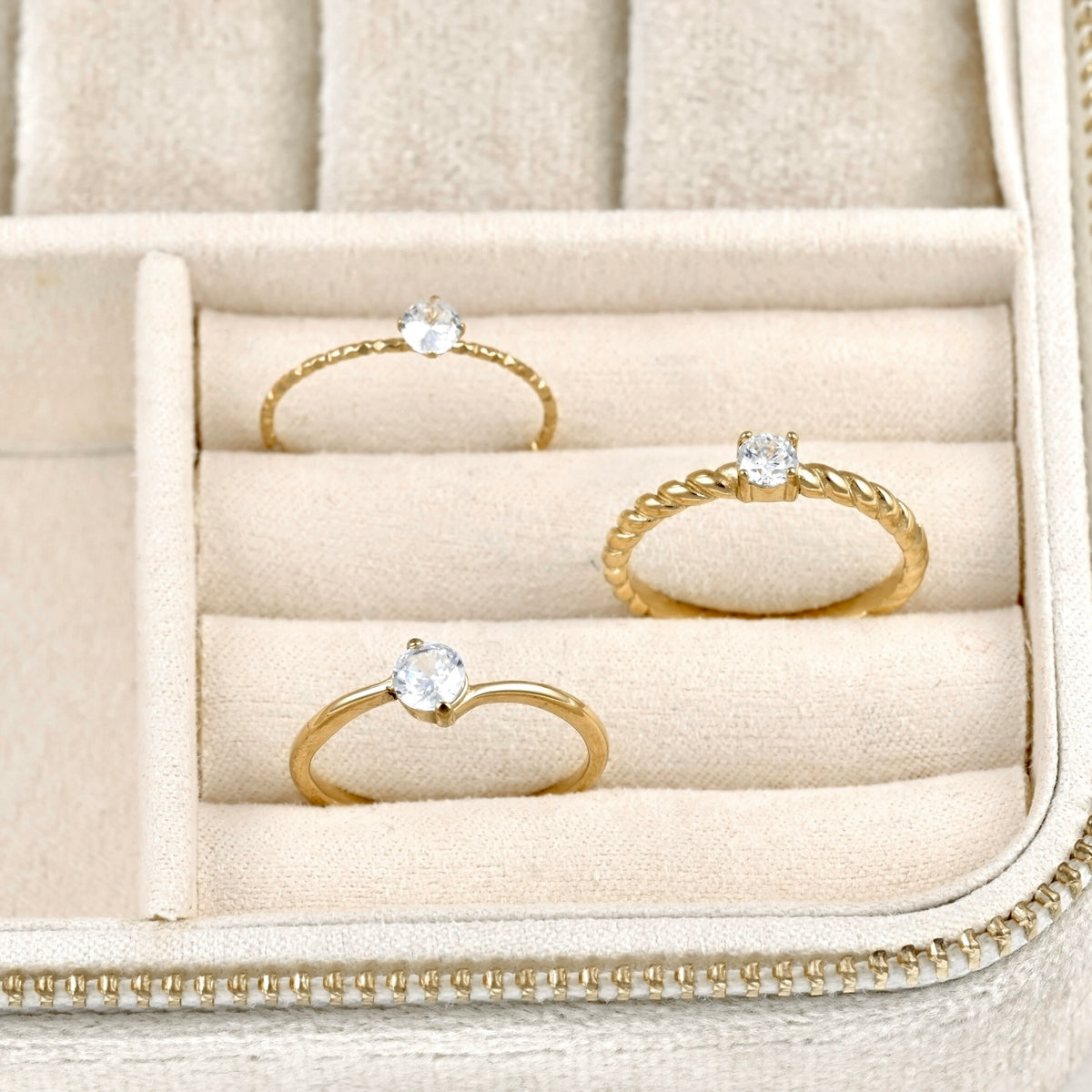 Three gold rings with clear gemstones displayed in a beige jewelry box.