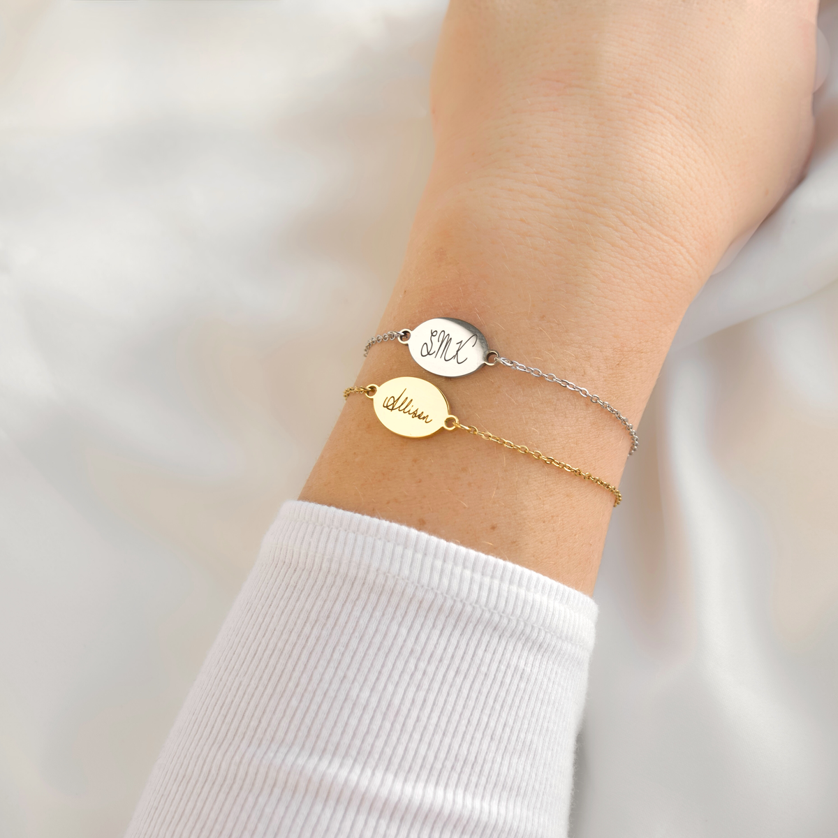 A close-up of a person's wrist wearing two delicate bracelets: a silver chain bracelet with a round engraved disc and a gold chain bracelet with a matching oval engraved disc, both featuring personalized script initials.