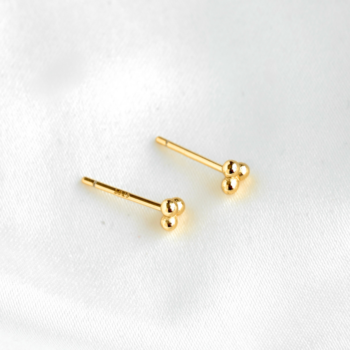 A pair of gold stud earrings with small spherical bead details at the end, displayed on a soft white fabric background.