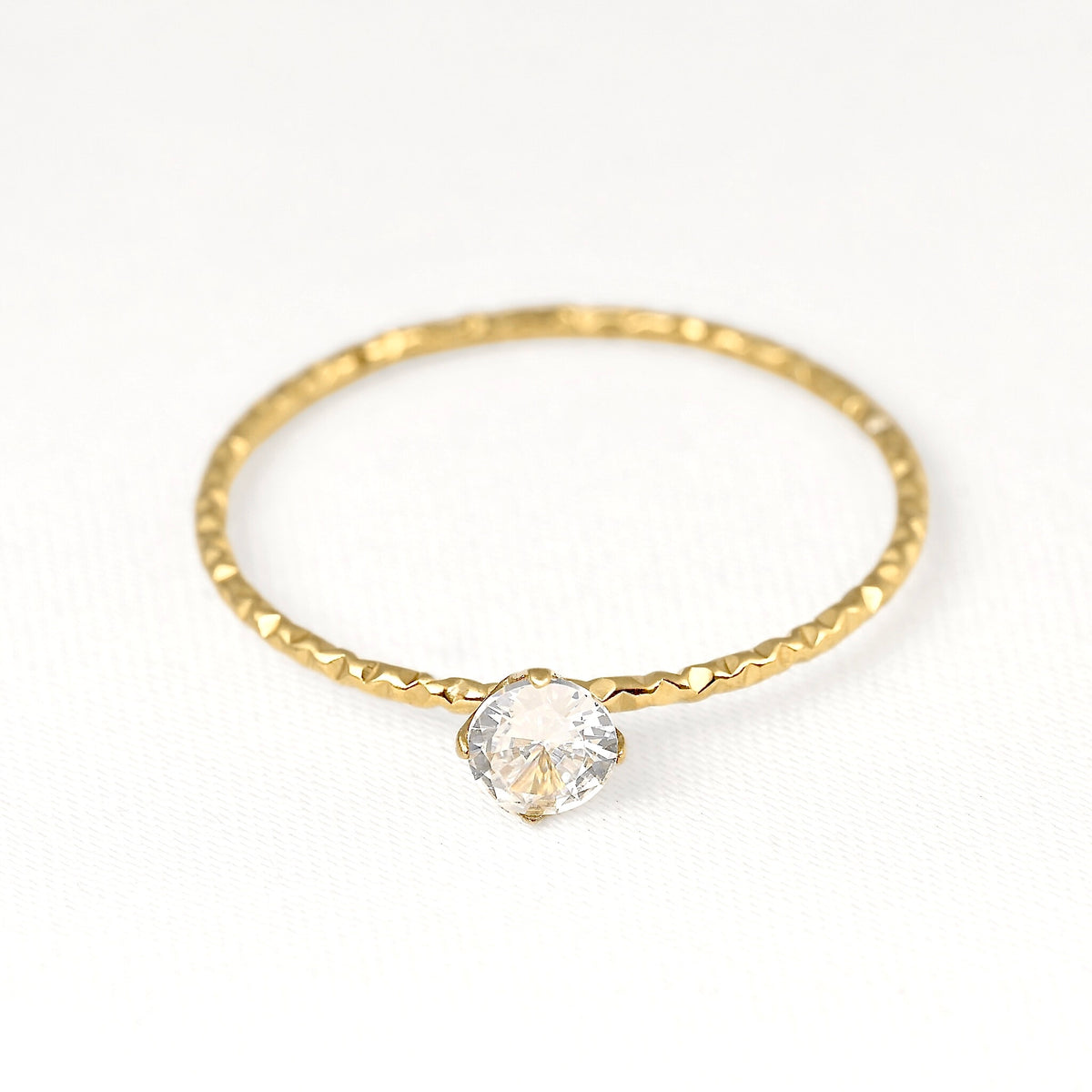 A delicate gold ring with a textured, hammered band and a single clear faceted gemstone in a prong setting, shown on a clean white background.