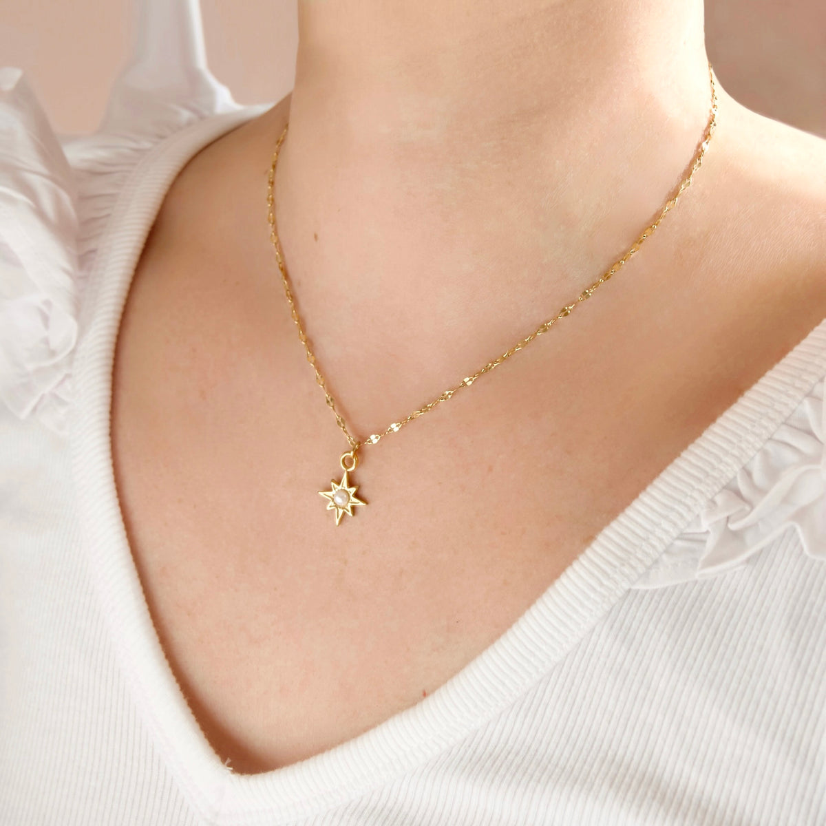 Angled close-up of a person wearing a delicate gold shimmering chain necklace with a small gold north star pendant featuring a central pearl, styled with a white ribbed V-neck top.