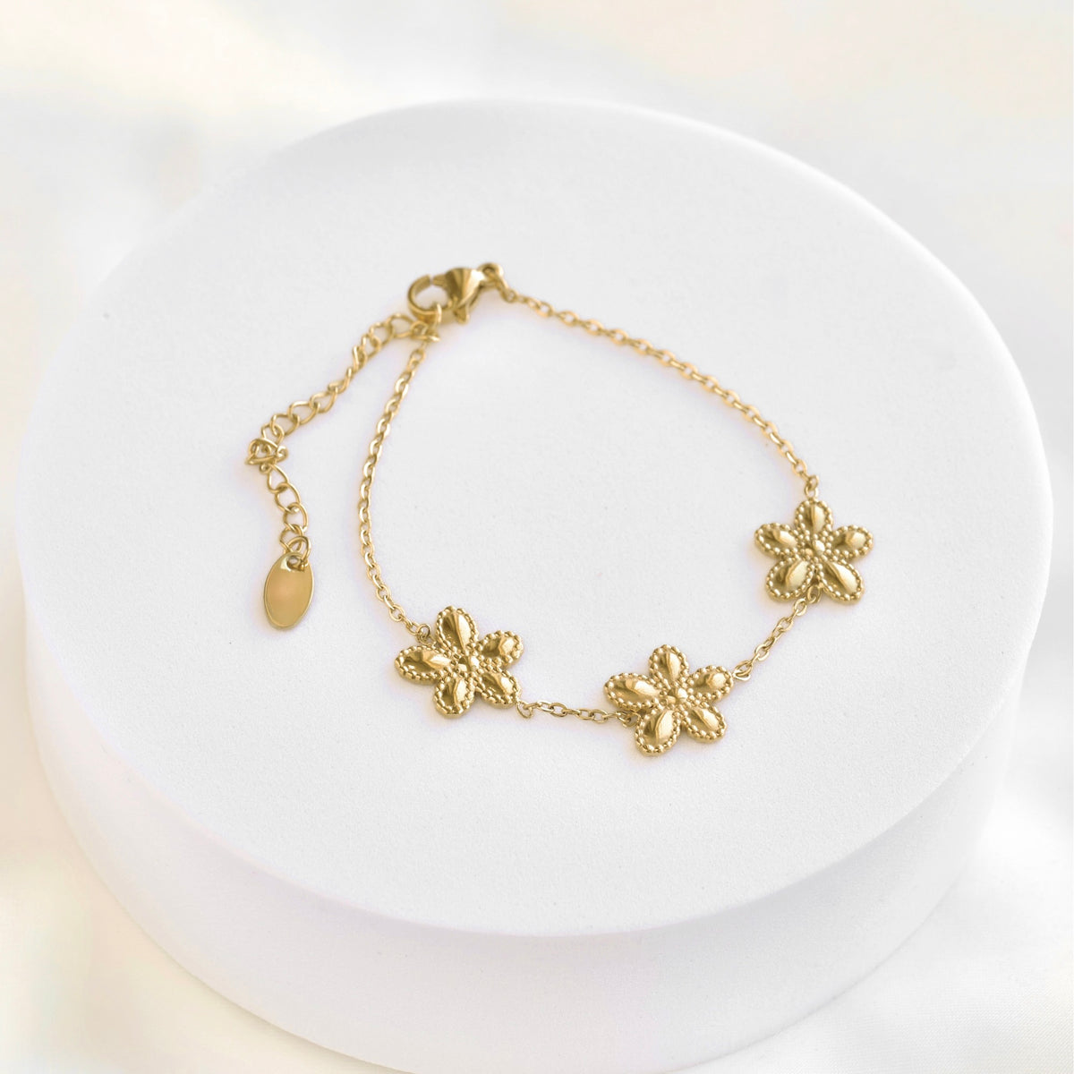 A delicate gold chain bracelet with three textured flower charms and an adjustable clasp, displayed on a round white pedestal.