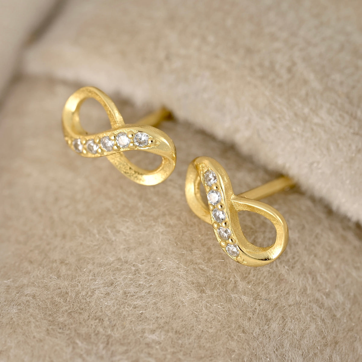 A pair of gold infinity-shaped stud earrings with small clear stones set along the curves, displayed on a soft beige textured surface.