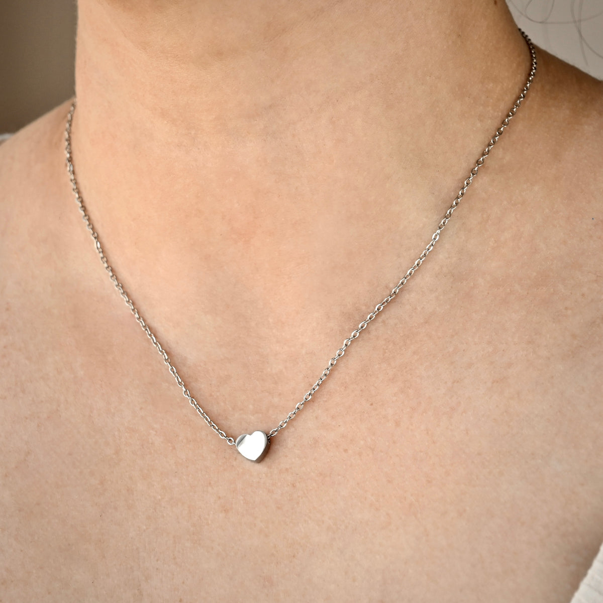 Silver necklace with a tiny heart pendant on a person's neck against a neutral background