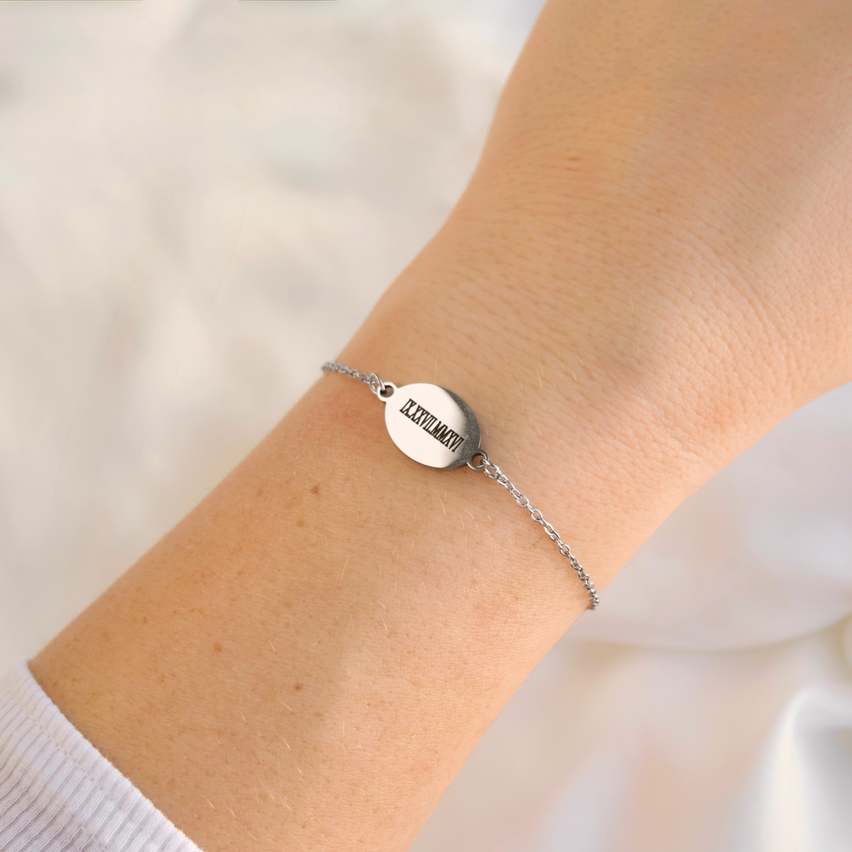 A delicate silver chain bracelet with a small oval disc engraved with the word 'LOVE' in black capital letters, worn on a person's wrist against a soft white background.