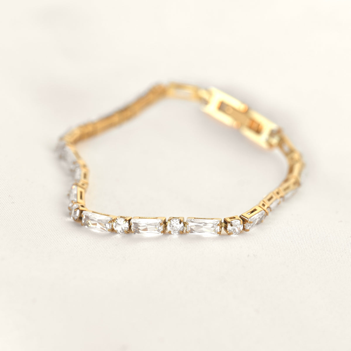 A yellow gold tennis bracelet featuring baguette and round cut clear crystals, displayed on a soft, light beige fabric background.