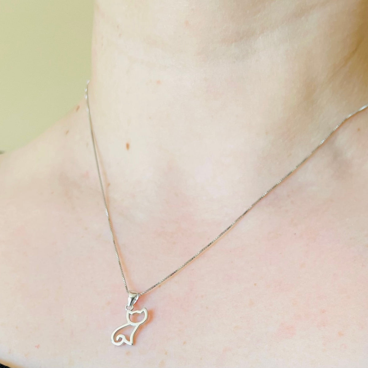 Silver necklace with sitting cat silhouette pendant on a person's neck against a neutral background