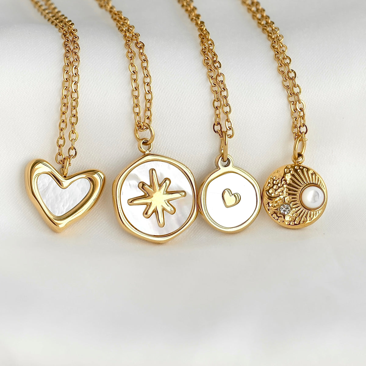 Four gold pendant necklaces with white inlay details, displayed on a soft white background. The pendants include a heart, a starburst, a small heart, and a textured sunburst with a pearl.