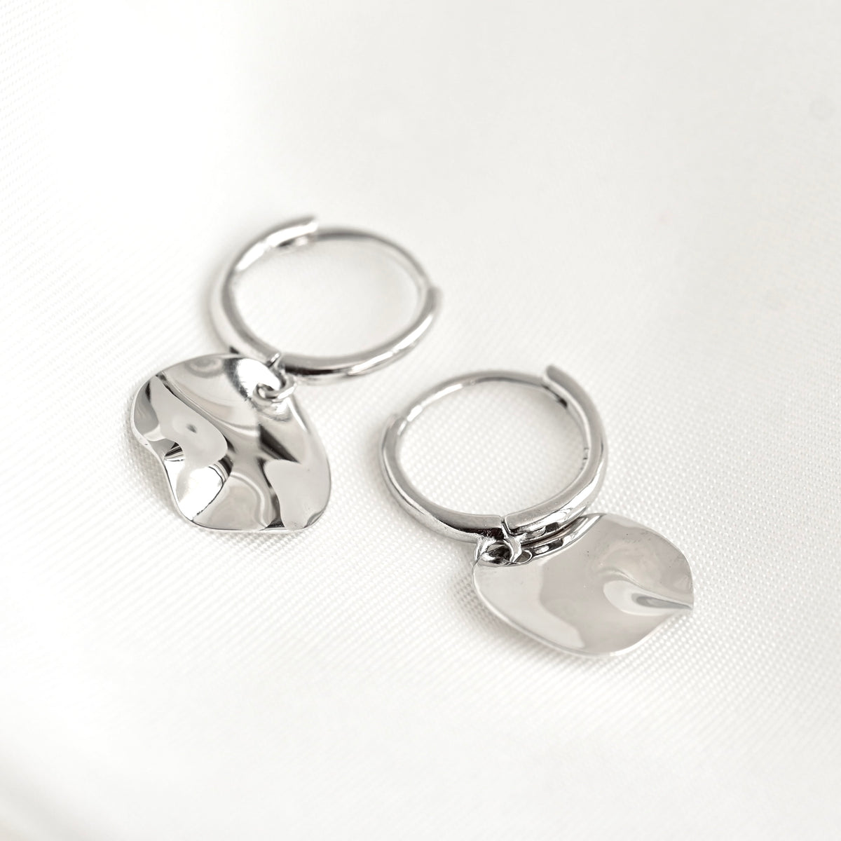 A pair of polished silver hoop earrings with irregular, organic-shaped dangling charms, displayed on a clean white textured fabric background.