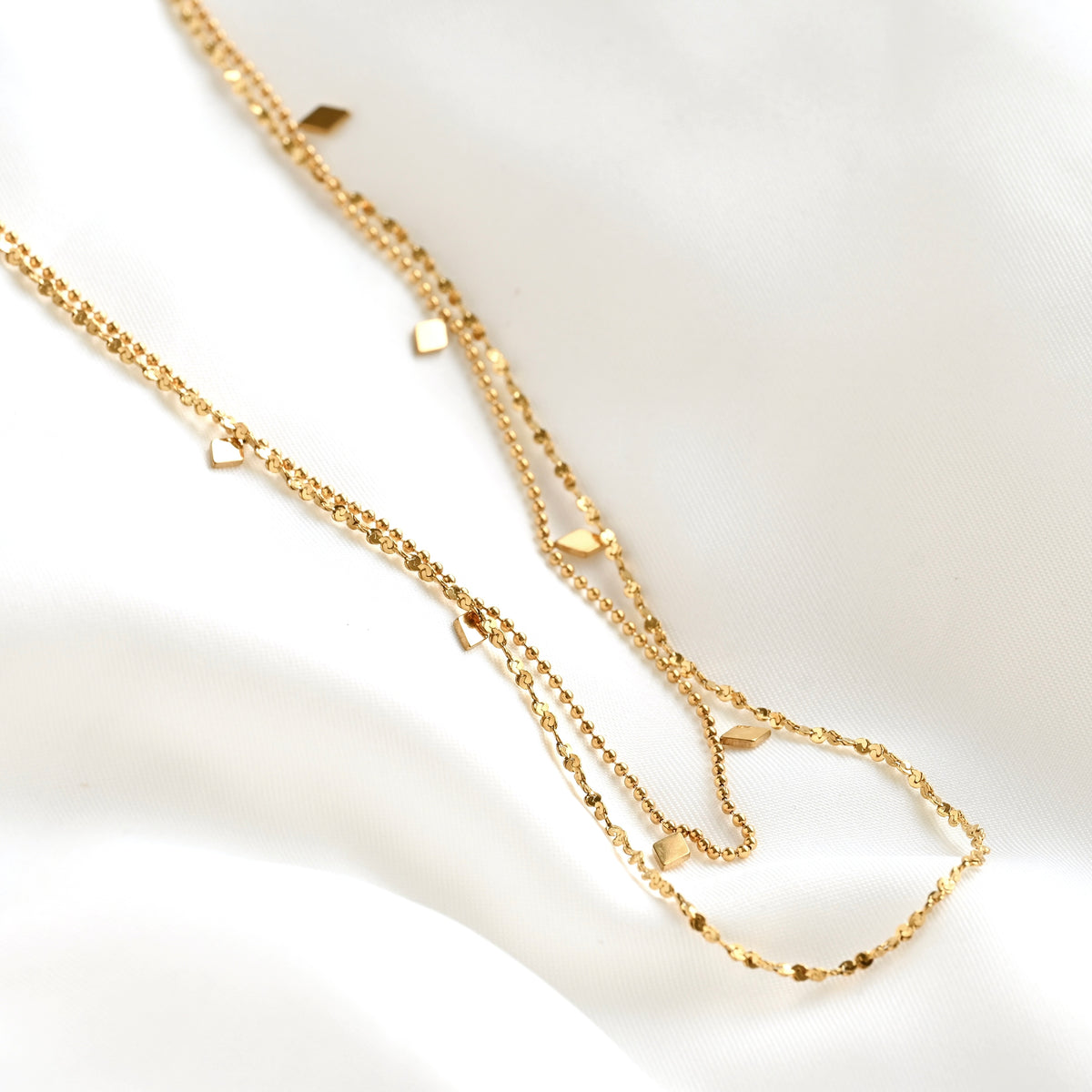 A delicate gold double-layer necklace with small diamond-shaped charms, laid out on a soft white fabric background.