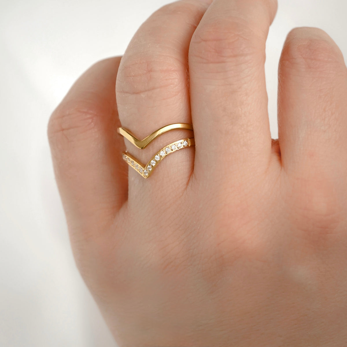A close-up of a hand wearing a gold chevron ring with one flat gold chevron and one pavé-set chevron, photographed against a soft light gray background.