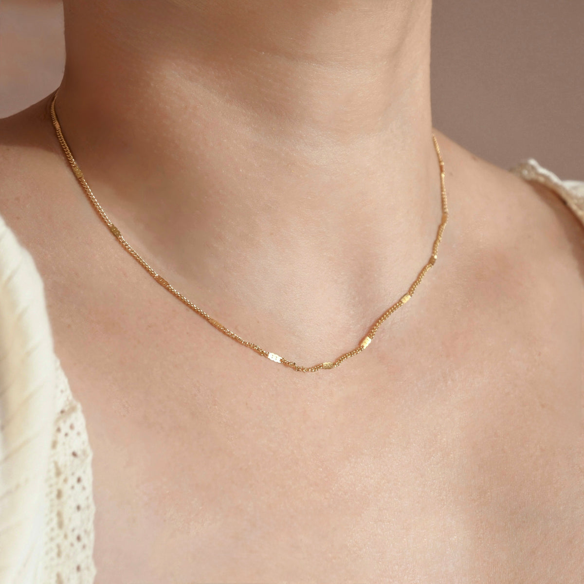 A delicate gold chain necklace with small flat rectangular links, worn on a model's neck over a cream-colored lace top.