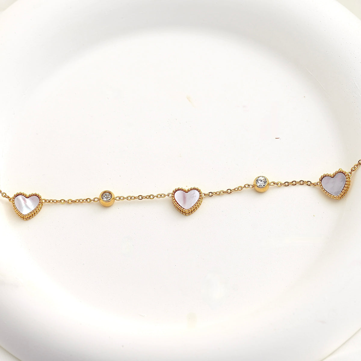 A delicate gold chain bracelet featuring three heart-shaped mother-of-pearl pendants and two small clear crystal beads, displayed on a white ceramic plate.