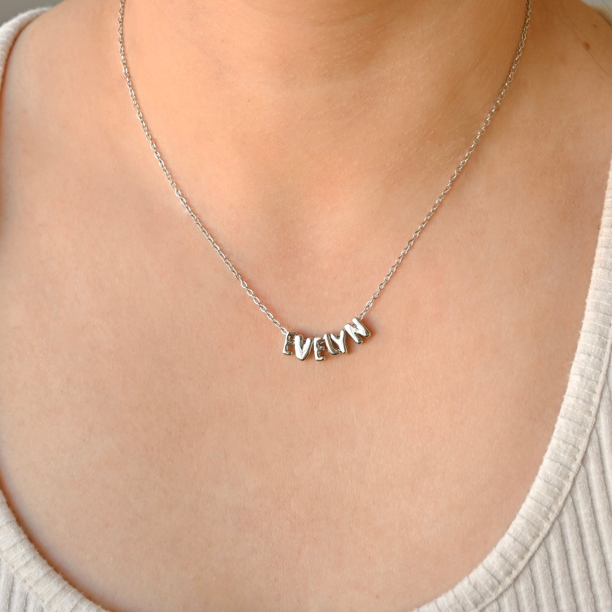 A close-up of a person wearing a delicate silver chain necklace with a personalized nameplate pendant spelling 'EvELYN' made with individual bubble initial charms