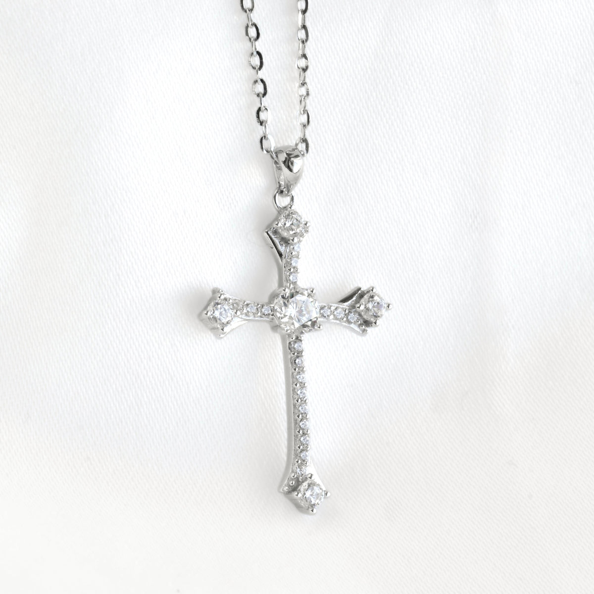 A silver cross pendant necklace adorned with clear gemstones, hanging from a silver chain against a white fabric background.