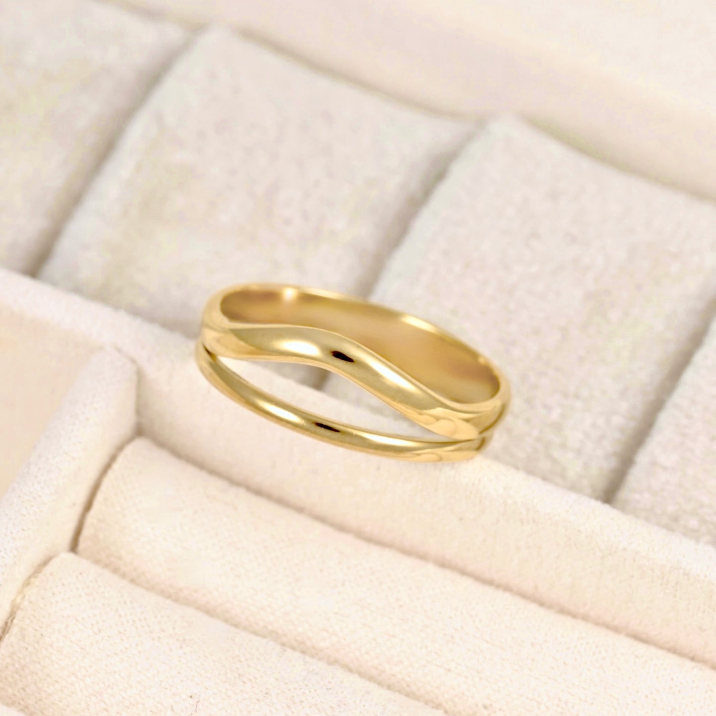 A polished gold ring with a wavy, double-band design resting on a textured cream-colored jewelry display box.
