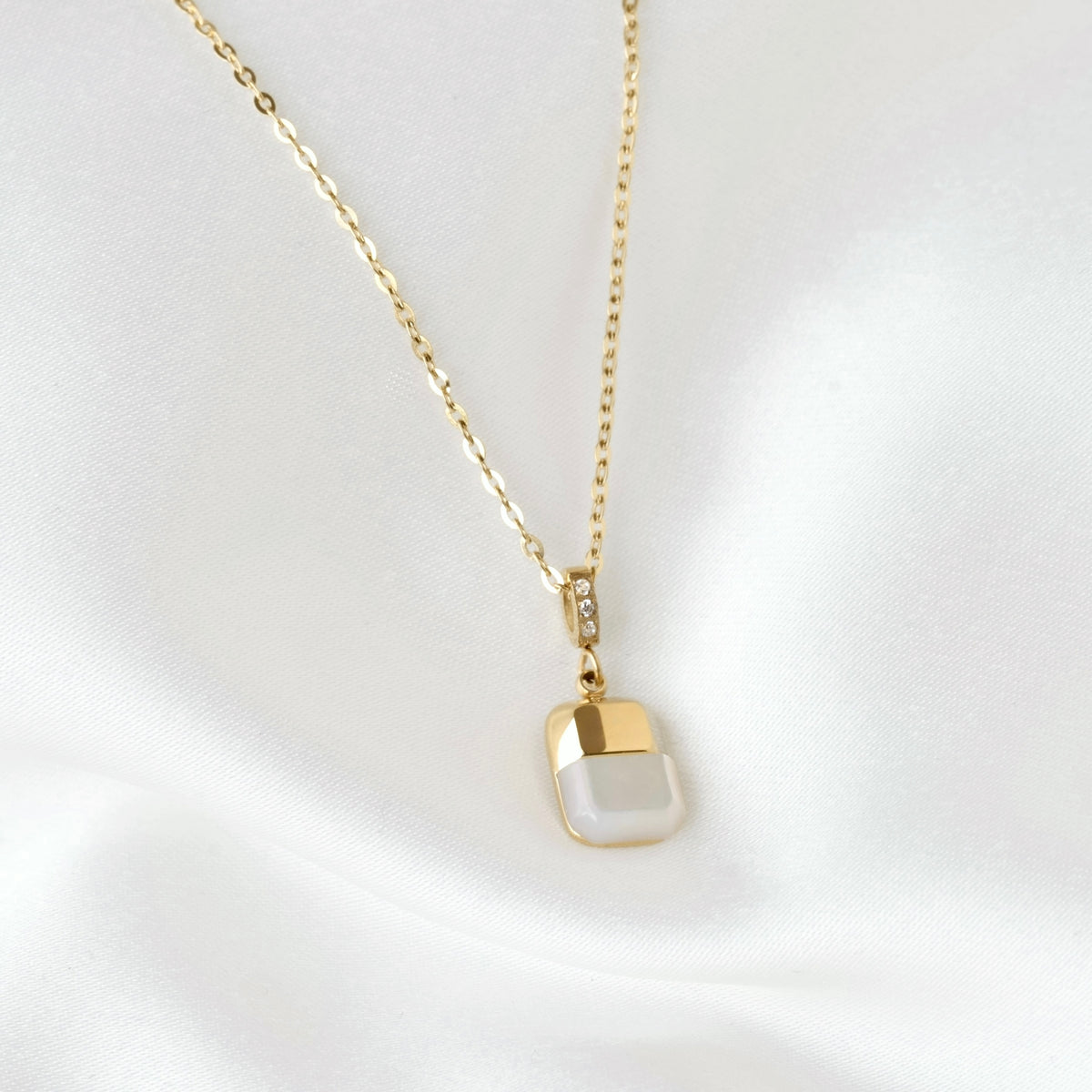 A delicate gold pearl necklace with a small rectangular pendant, featuring a white pearl inlay on the bottom half and a polished gold top half, displayed on a soft white fabric background.