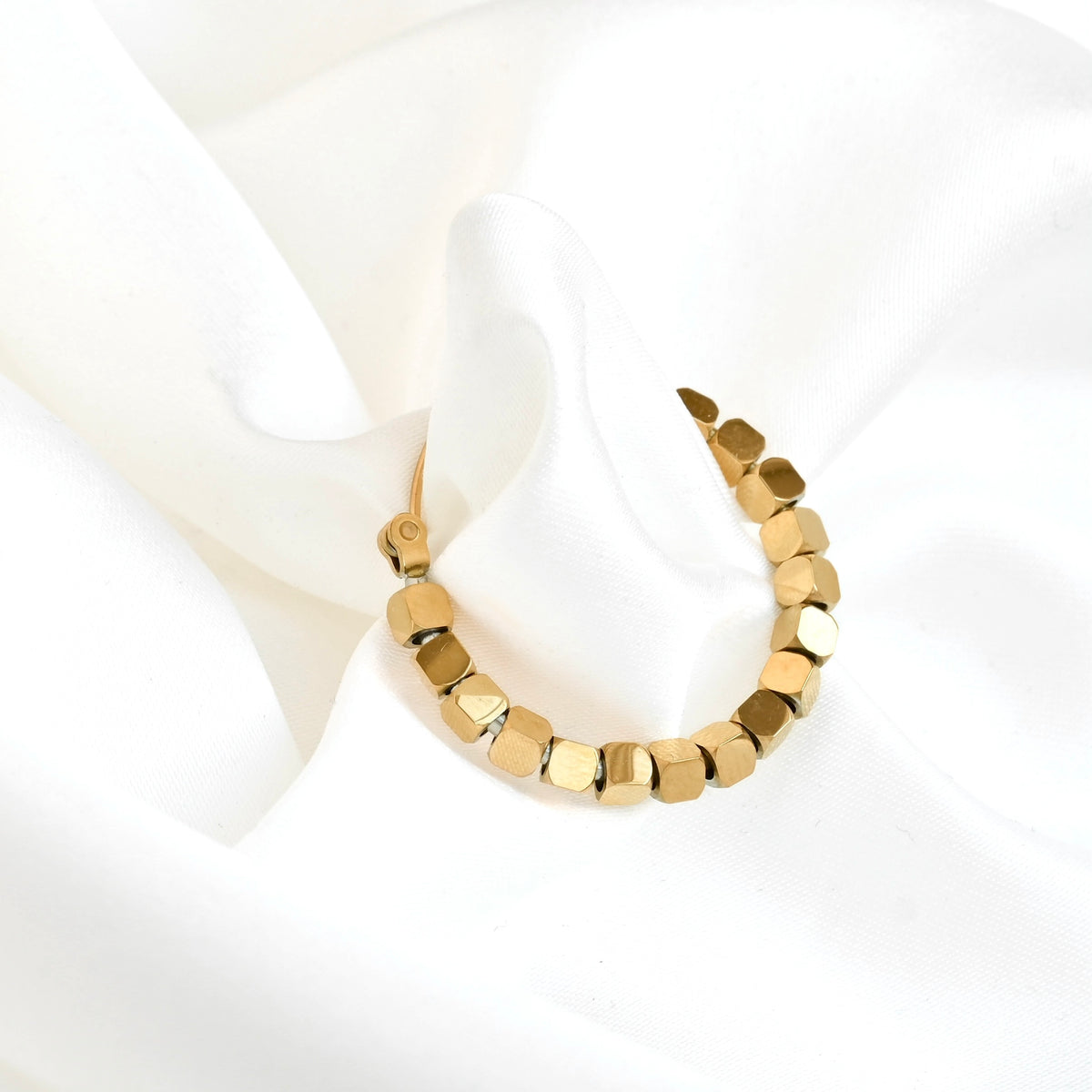 A gold hoop earring with faceted, geometric cube beads, displayed on a soft, draped white fabric background.
