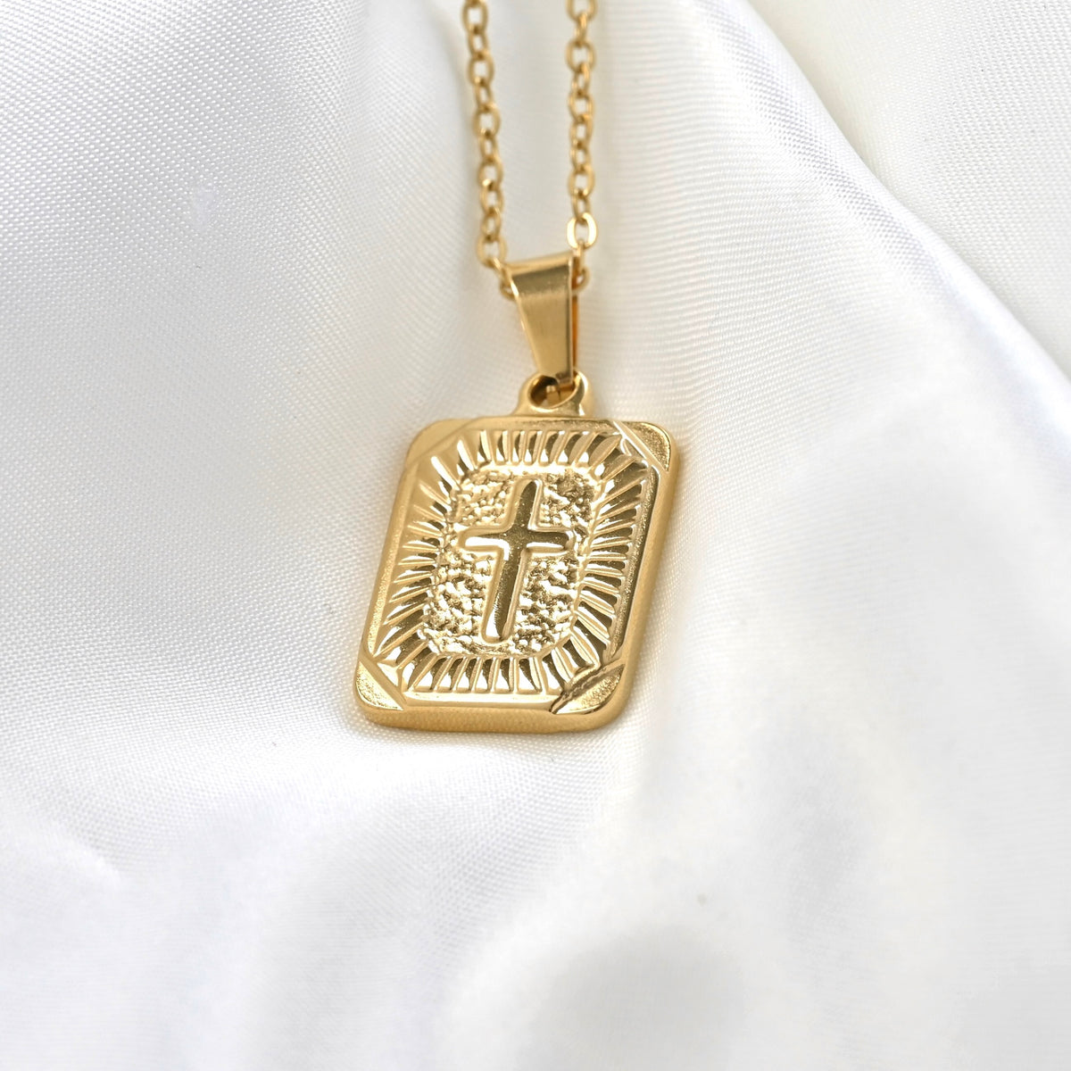 A gold-toned rectangular pendant necklace with an embossed cross and decorative border, hanging from a matching chain on a white satin fabric background.