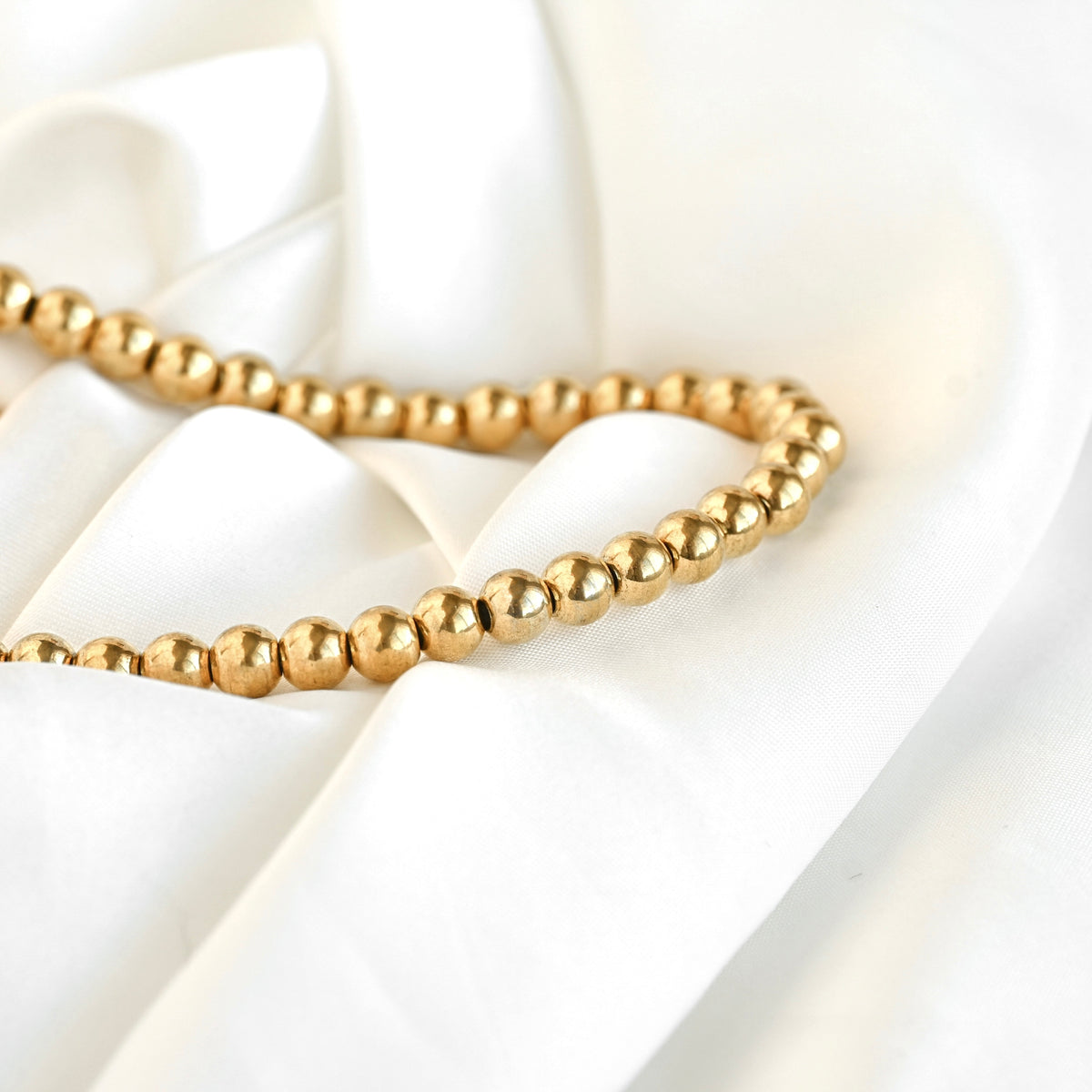A strand of small, polished gold-colored beads draped elegantly over soft, white satin fabric.