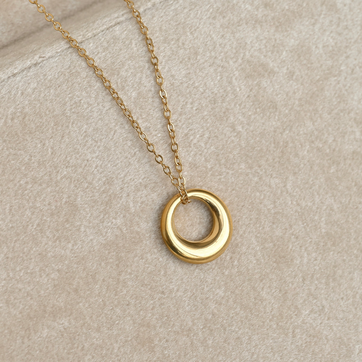 A gold chain necklace with a thick ring pendant, displayed on a textured beige fabric background.