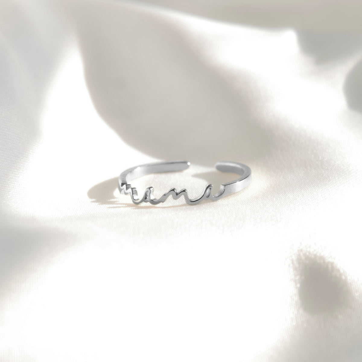 A silver open ring with a wavy, flowing 'mama' script design across the front, resting on a softly lit white fabric background.