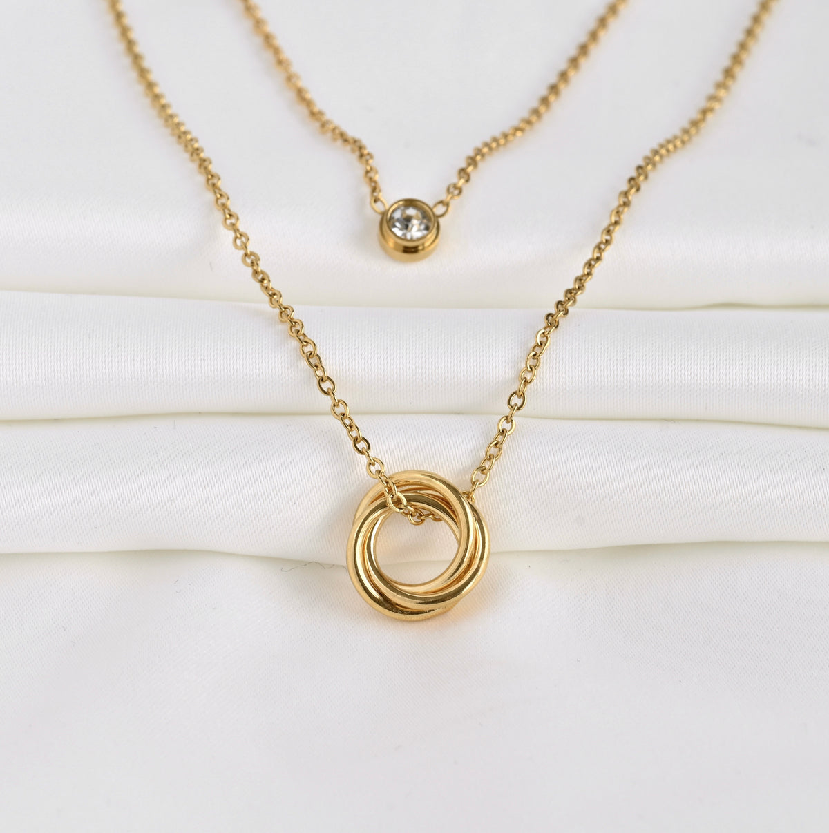 Wide angled front view of two layered gold necklace set with interlocking rings and a bezel charm on a white background