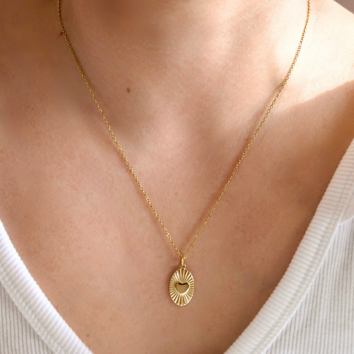 A close-up of a person's neck and chest wearing a delicate gold chain necklace with a small oval pendant featuring a sunburst design and a central heart emboss, styled with a white ribbed V-neck top.