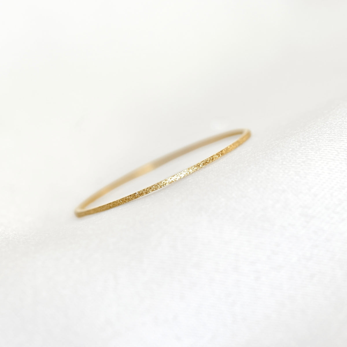 A delicate, thin gold ring with a textured, glittery finish, resting on a soft white fabric surface.