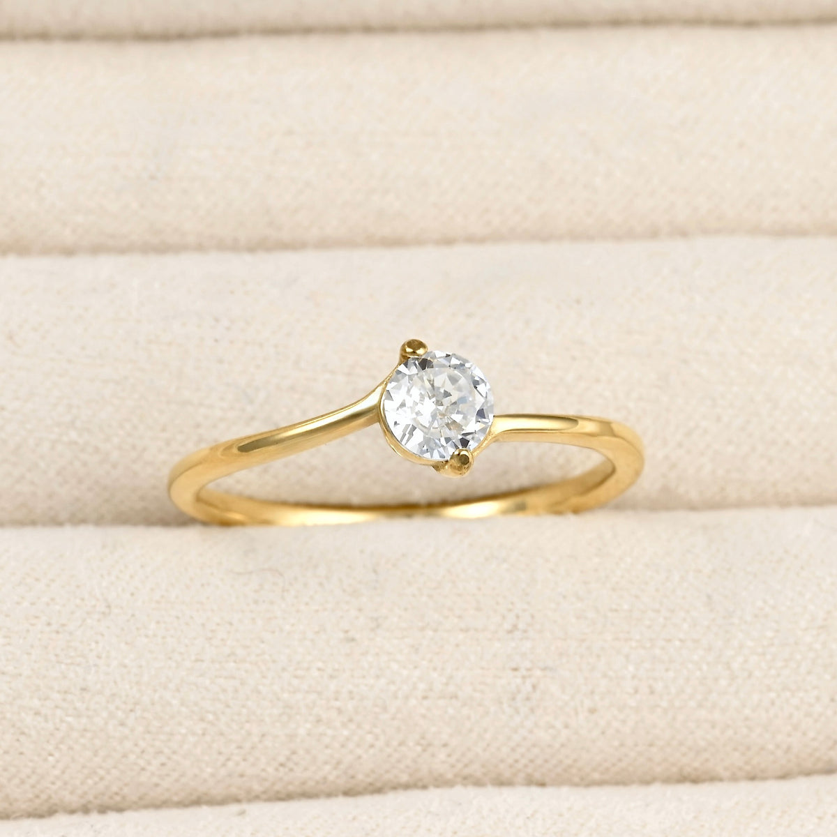 A yellow gold solitaire ring with a round-cut clear gemstone, displayed on a textured beige fabric ring box.