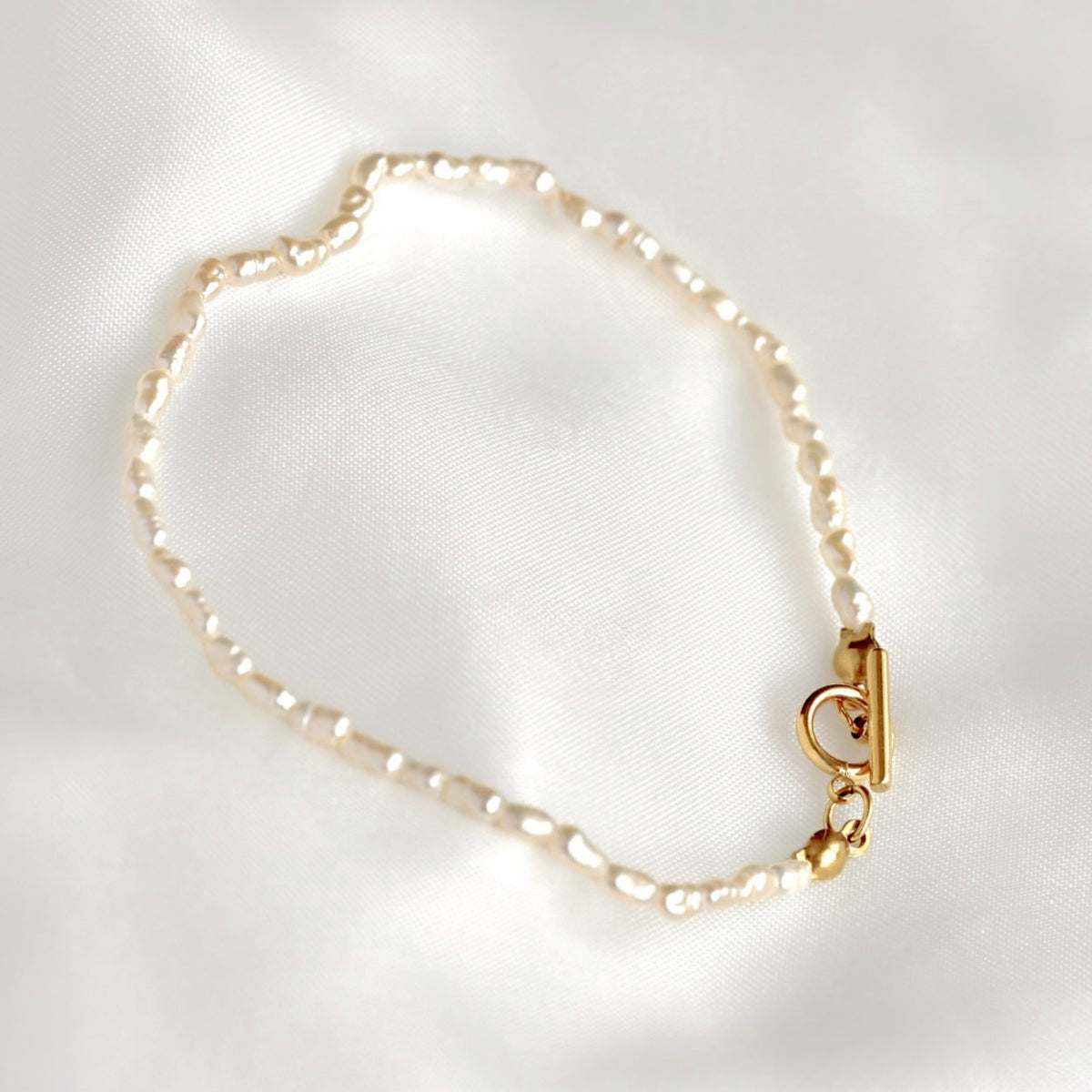 A delicate bracelet made of small irregular white rice pearls linked with a gold-tone toggle clasp, displayed on a soft white satin fabric background.