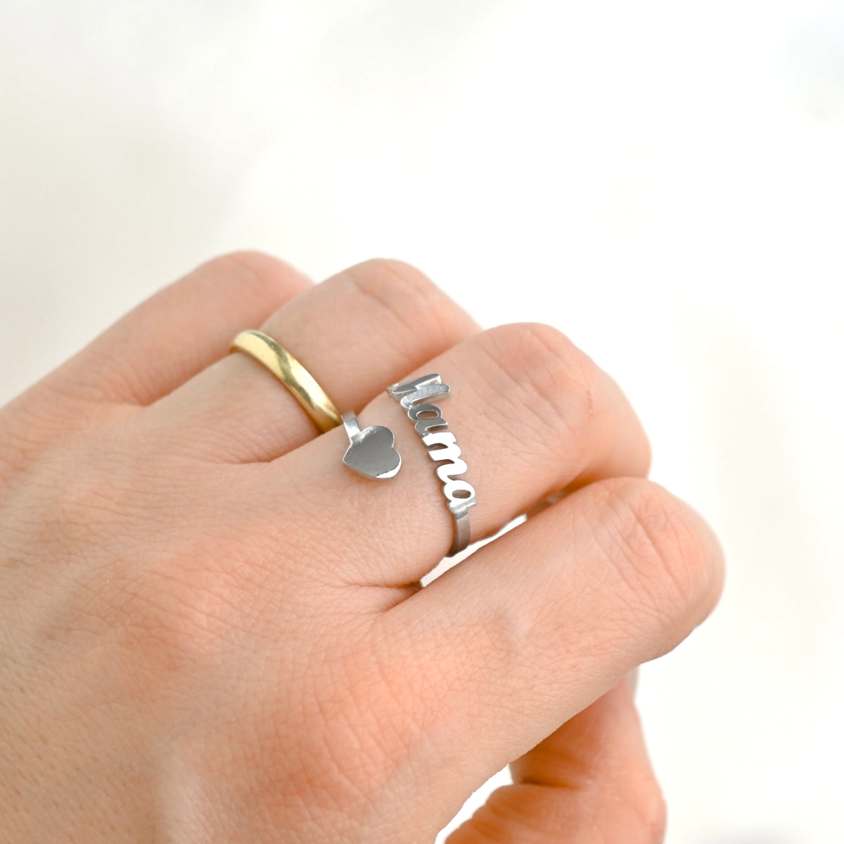 A close-up of a hand wearing two rings: a simple gold band and a silver mama heart