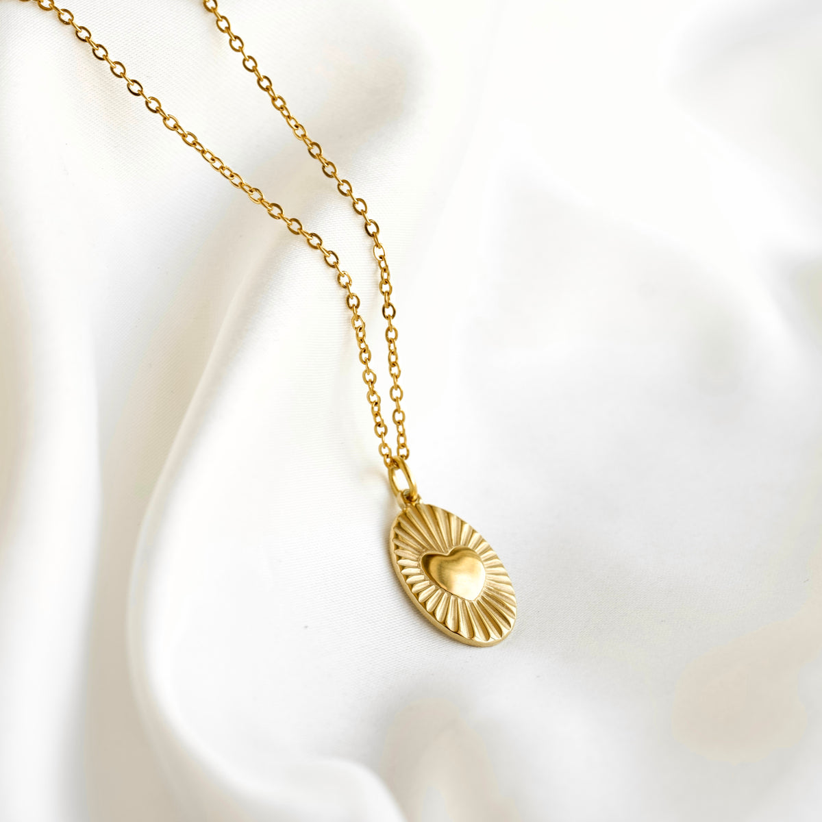 A gold chain necklace with an oval pendant featuring a sunburst design and a small heart in the center, displayed on a soft white fabric background.
