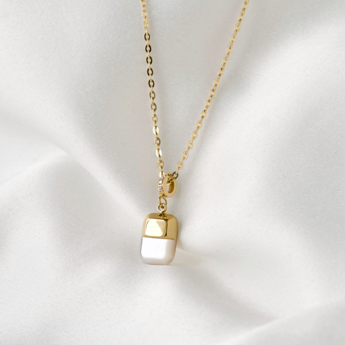 A gold chain necklace with a rectangular pendant featuring a glossy pearl bottom half and a polished gold top half, displayed on a soft white satin fabric background.