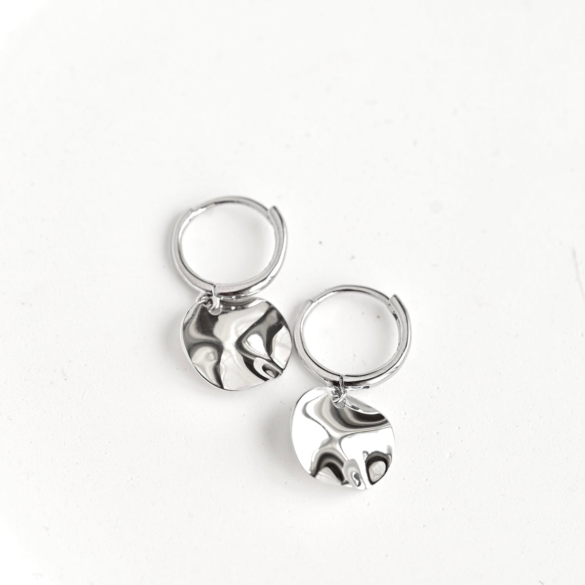 A pair of silver huggie hoop earrings with flat, textured circular dangles, displayed on a clean white background.