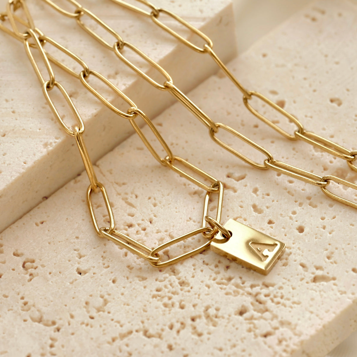 Gold paperclip necklace with a initial tag pendant on a textured beige surface