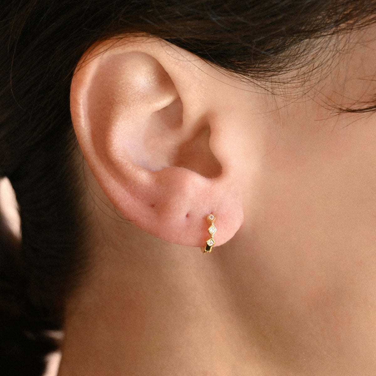 A close-up of a person's ear wearing a small gold huggie hoop earring with three tiny rhombus-shaped clear stones in a bezel setting
