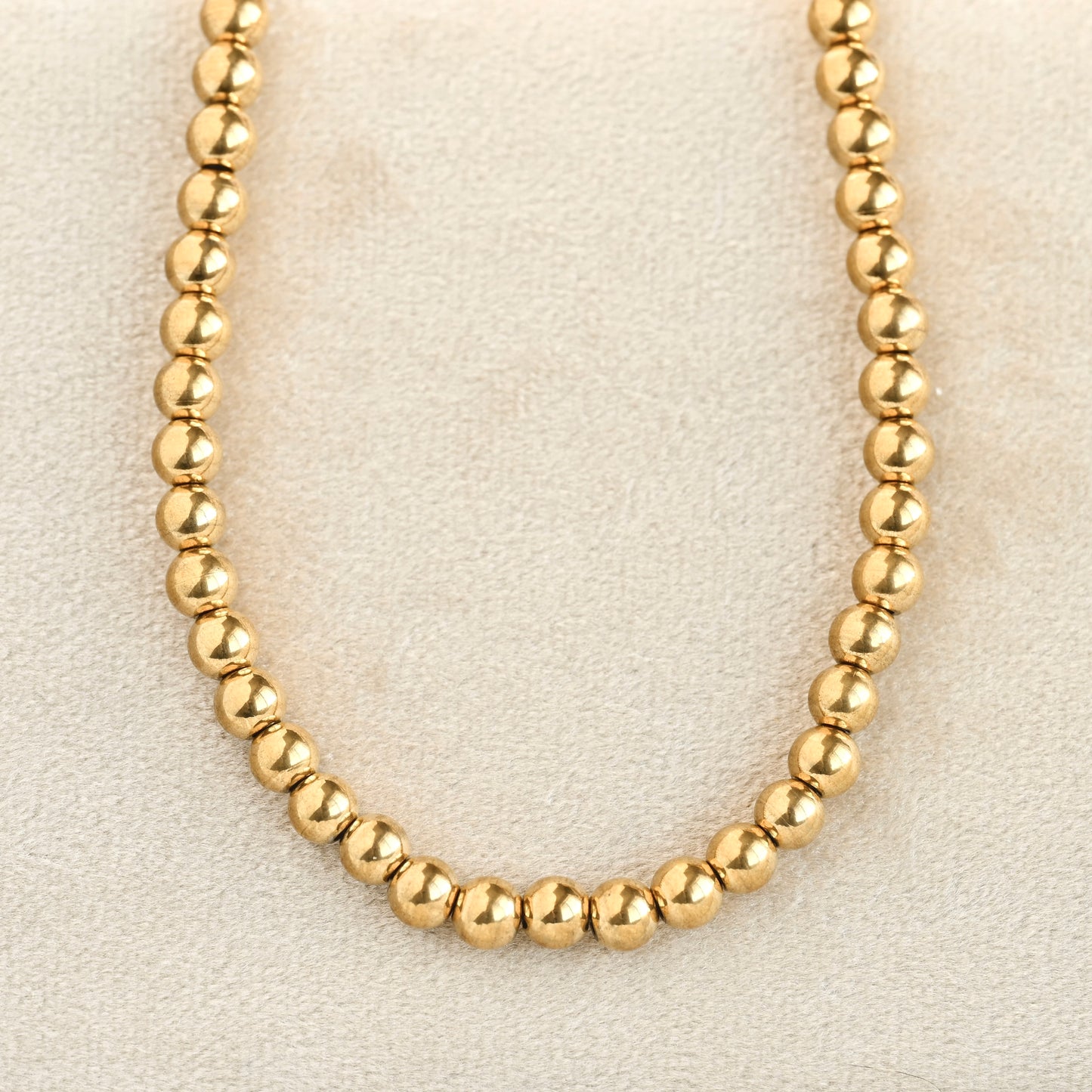 A gold beaded necklace made of small, polished round beads, displayed on a textured beige surface.