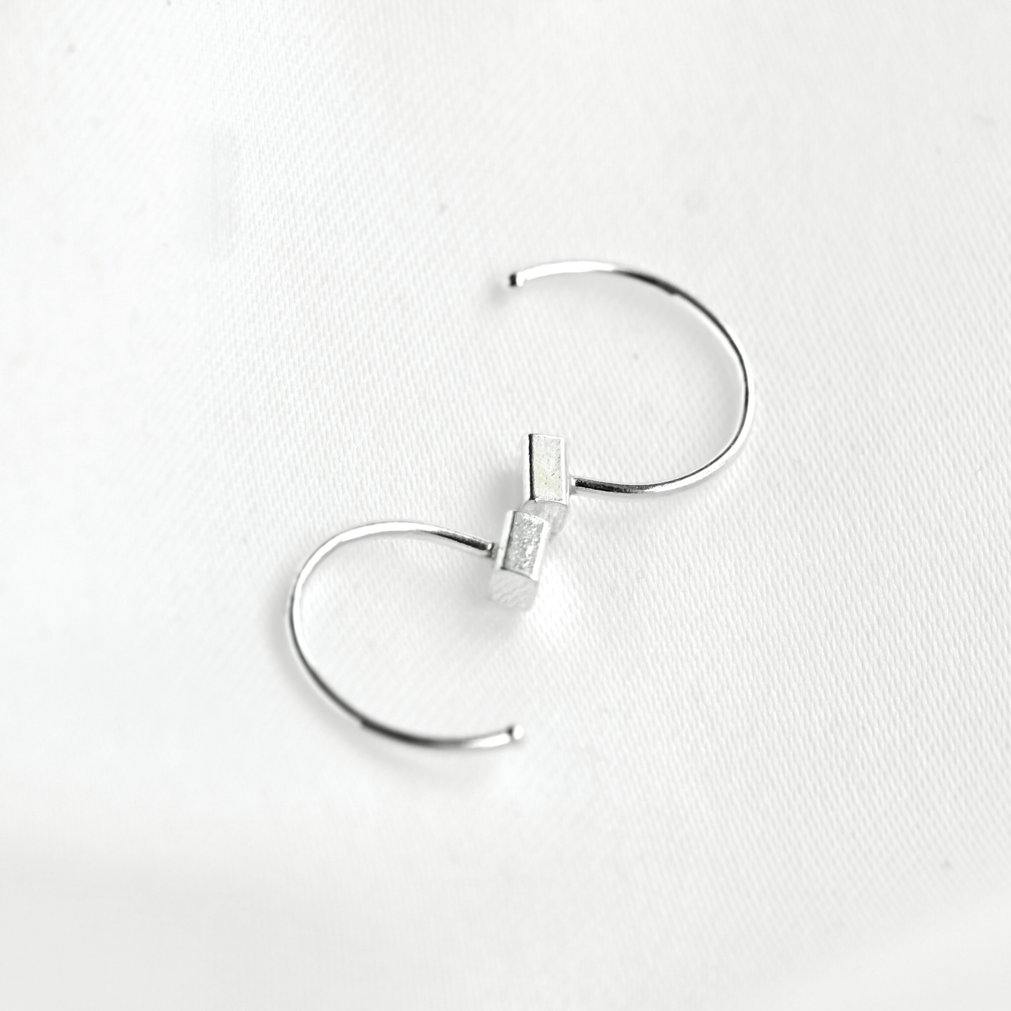 A pair of silver-toned open hoop earrings with a small rectangular bars on one end, displayed on a textured white surface.