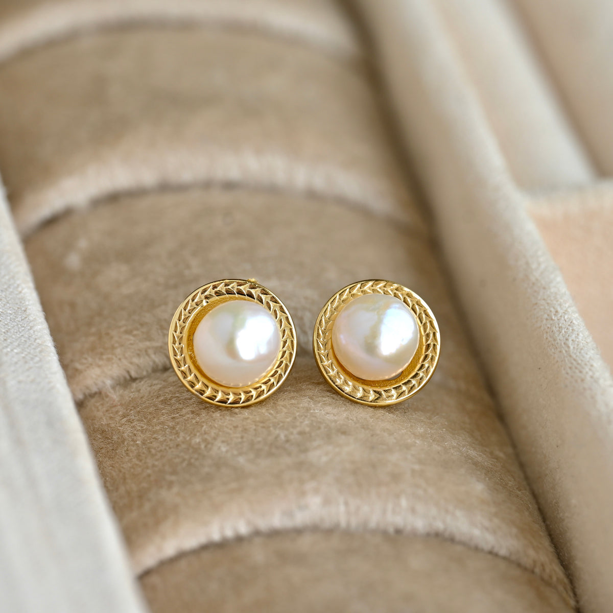 A pair of vintage style gold pearl stud earrings with round white pearls and a braided gold border, displayed on a beige velvet jewelry ring holder.